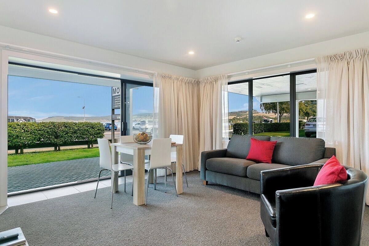 A modern living room with a grey sofa, a dining table with chairs, and large windows offering a view of the outdoors.