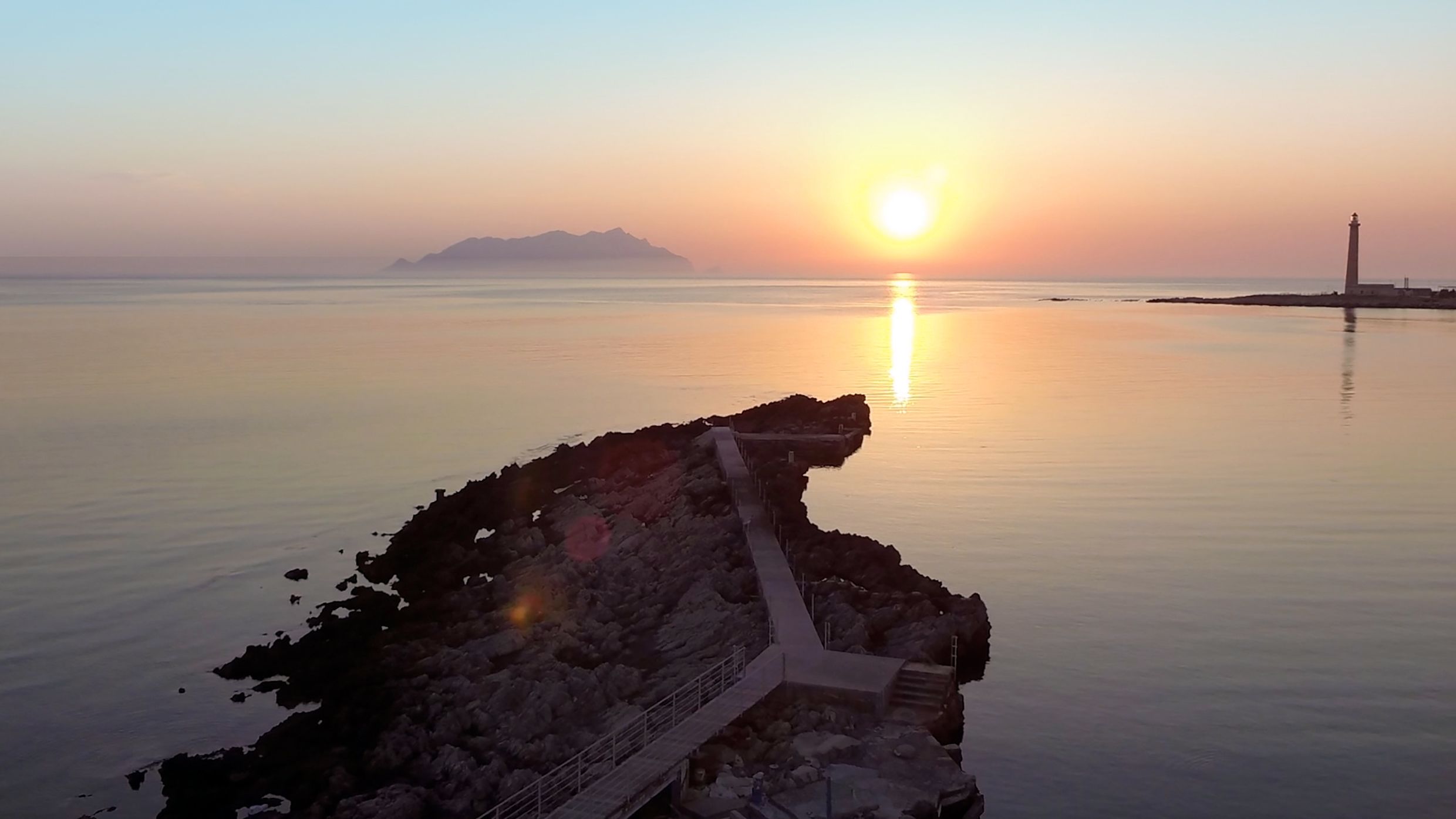 A coastal scene with a pier extending into calm waters, a distant mountain, and a lighthouse.