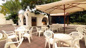 An outdoor patio with white plastic chairs and tables, a large umbrella, and a building with arches in the background. - Sabbie Bianche (Parghelia)