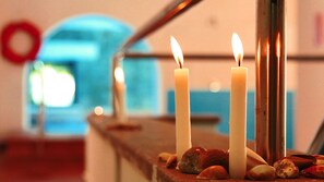 Three lit candles on a ledge with rocks and seashells, a metal railing, and a blurred background with a blue and orange hue. - Villaggio dei Pini (Sorso)