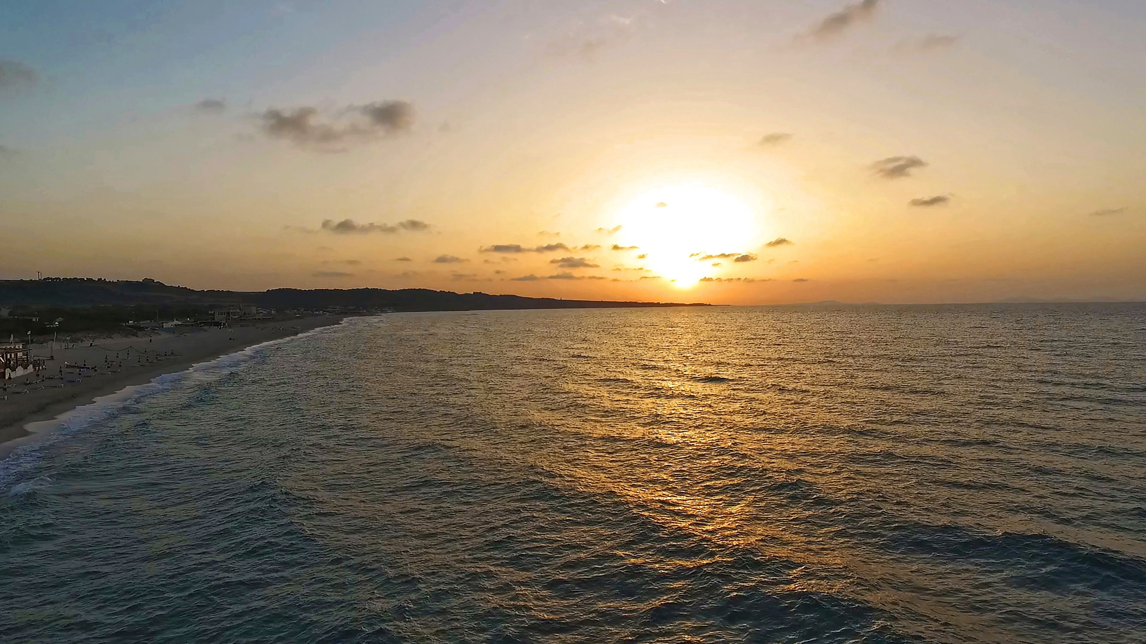 Uma praia com o pôr do sol no oceano, um cais à esquerda e uma paisagem montanhosa ao longe.