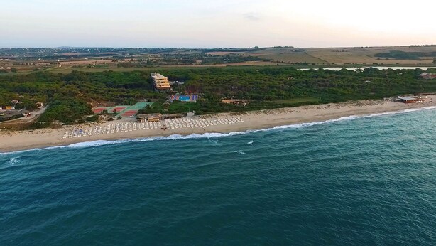An aerial view of a coastal area with a beach, a hotel with multiple floors, and surrounding greenery. - Villaggio dei Pini (Sorso)