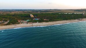An aerial view of a coastal area with a beach, a hotel with multiple floors, and surrounding greenery. - Villaggio dei Pini (Sorso)