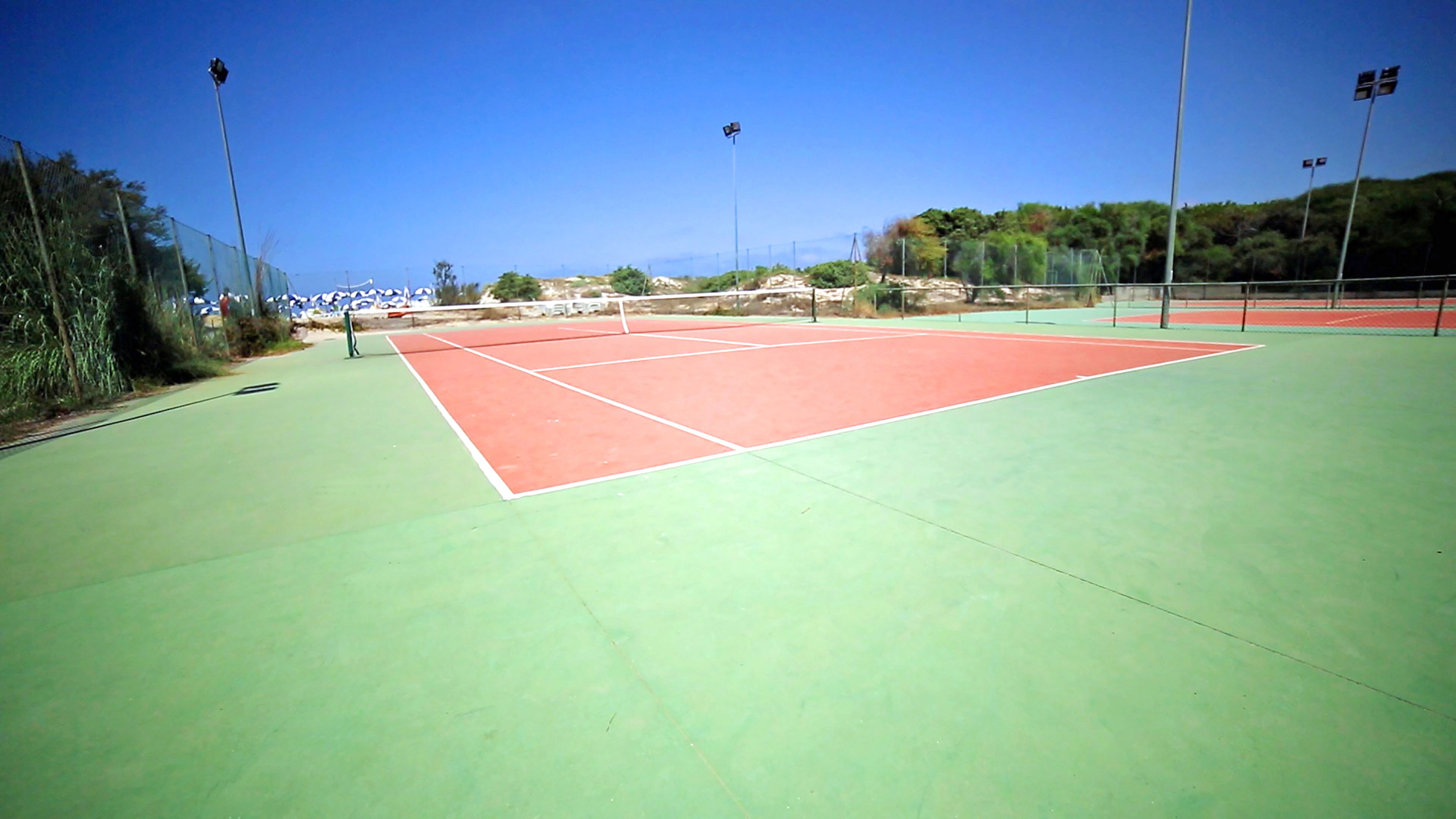 An outdoor tennis court with green and red surfaces, surrounded by fencing and trees.