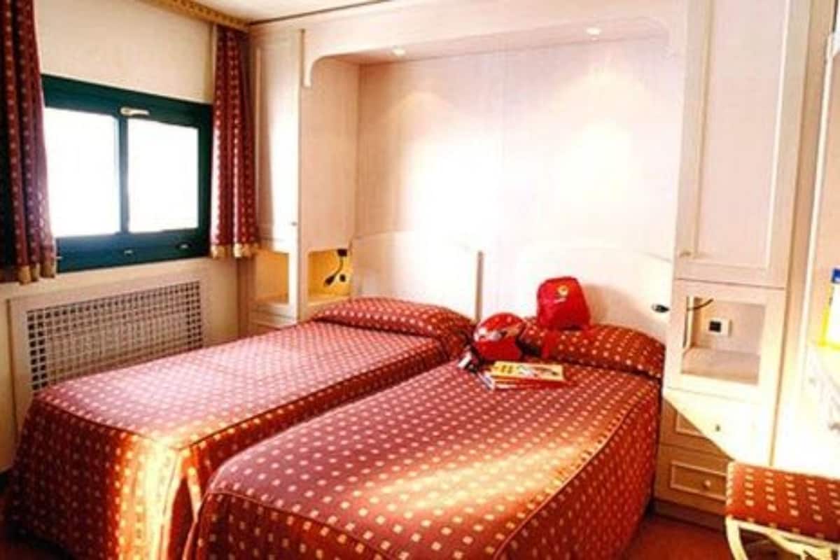 Una habitación de hotel con dos camas individuales, una mesita, una silla, una ventana con cortinas y un radiador.