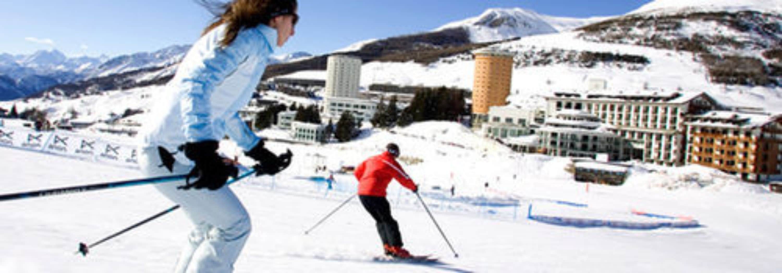 A snowy ski slope with skiers, buildings, and mountains in the background.