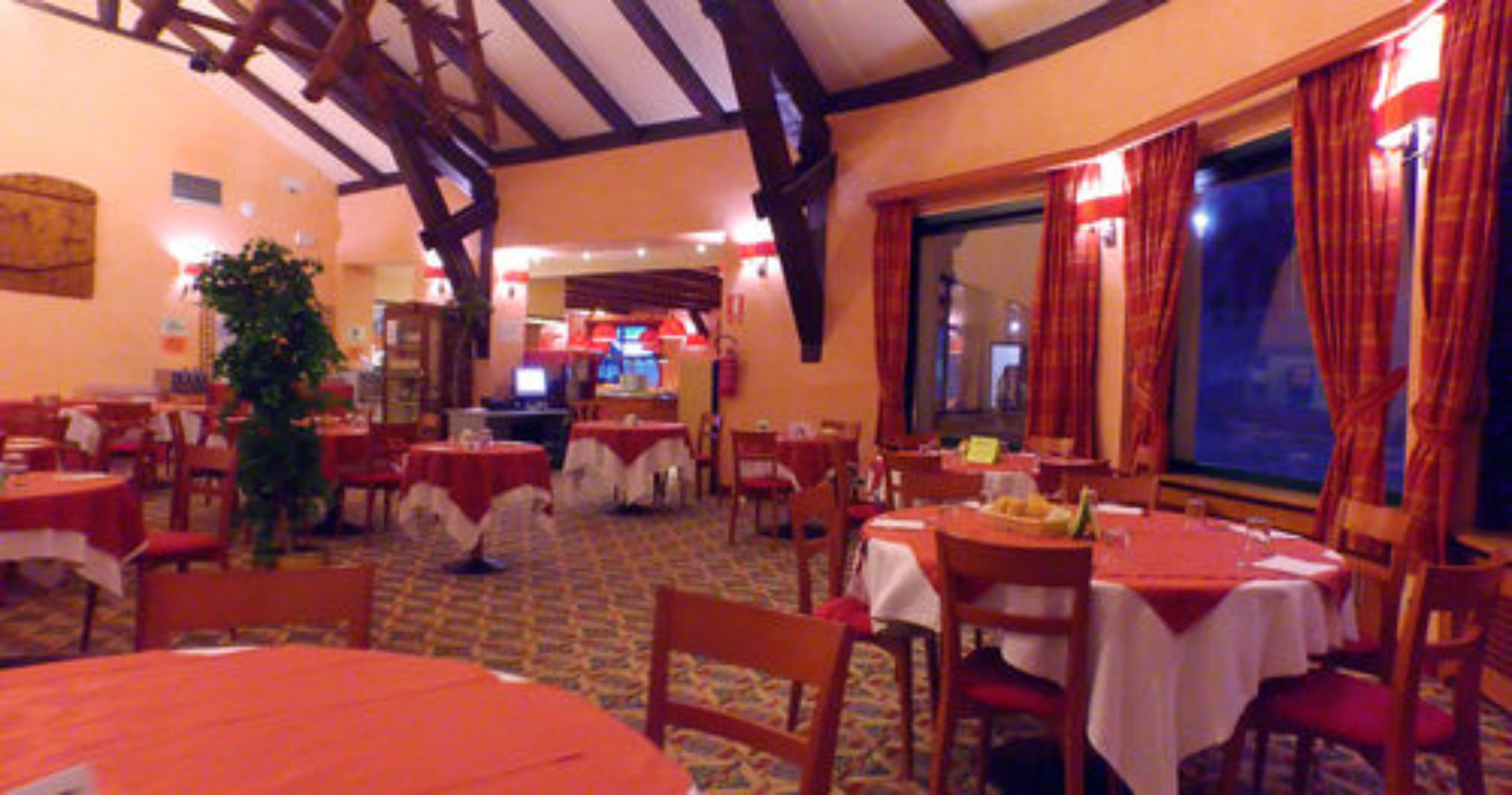 Ein Restaurant mit mehreren Tischen und Stühlen, Holzbalken an der Decke und einem großen Fenster mit Vorhängen.