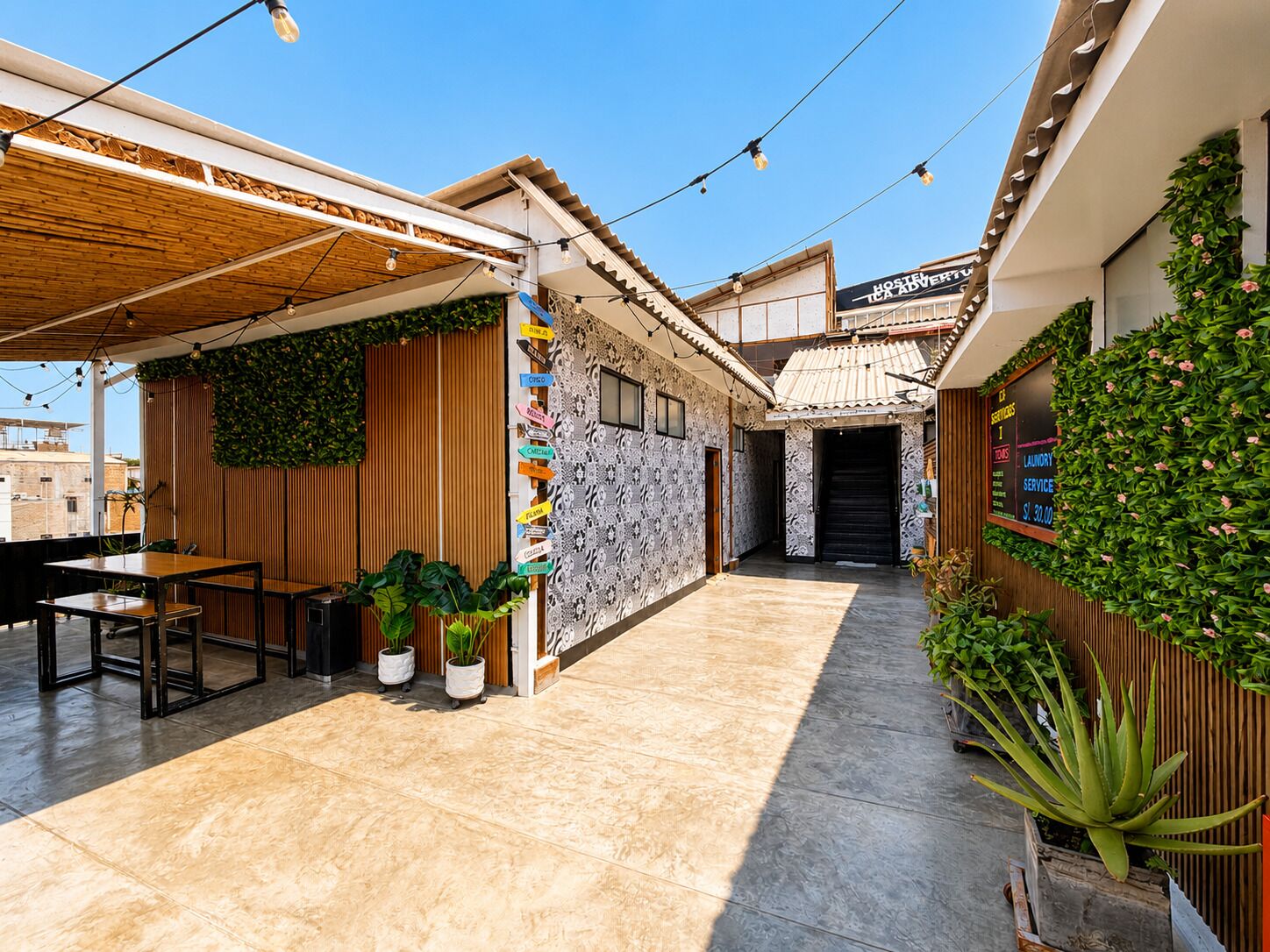Um pátio com pérgola de madeira, plantas em vasos, uma parede com placas de sinalização e um edifício com a placa 'Hostel La Aventura'.