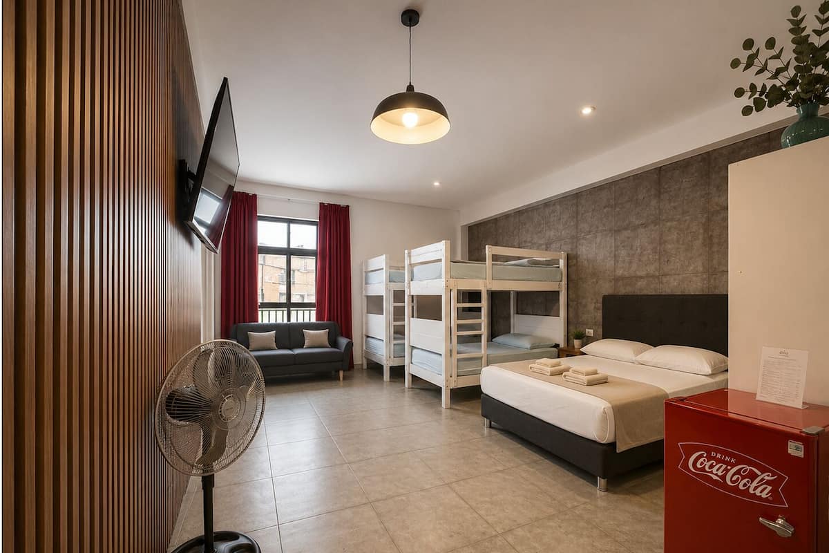 A hotel room with bunk beds, a sofa, a fan, a television, and a Coca-Cola refrigerator.