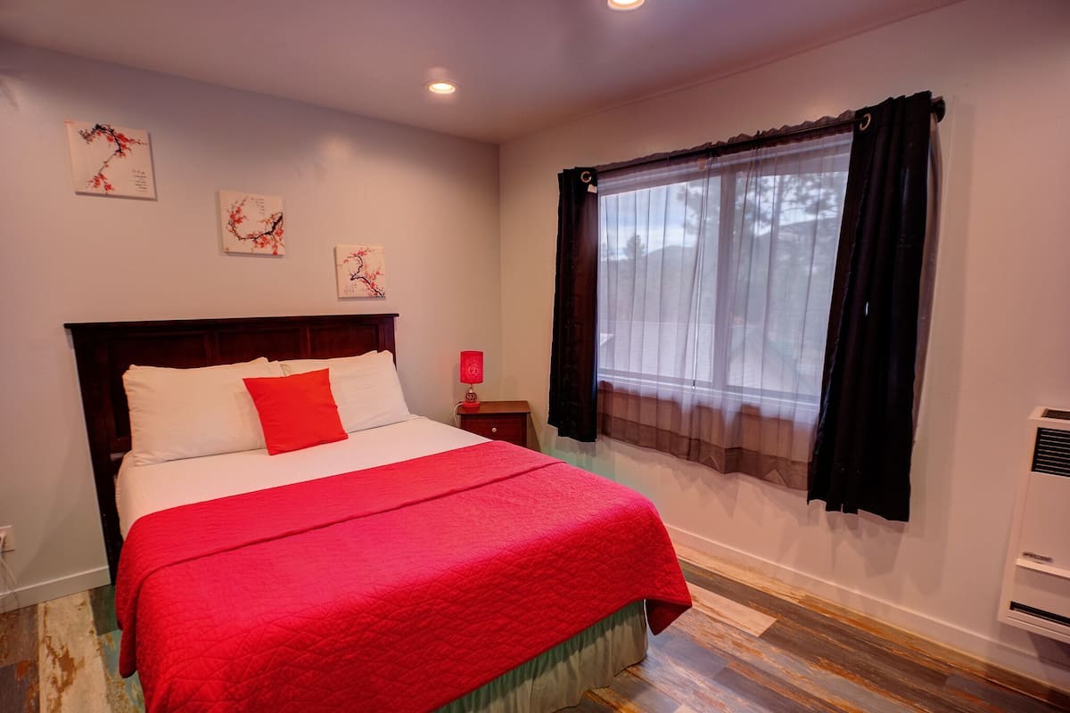 Una cama bien hecha con una colcha roja, una lámpara de noche y una obra de arte en la pared.