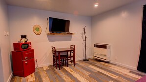 Una habitación con una televisión montada en la pared, un refrigerador rojo, una mesa de comedor con sillas, una lámpara de pie y un calentador blanco.