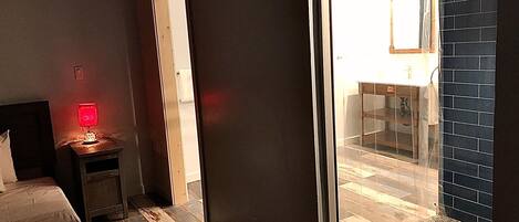 A hotel room with a bed, a nightstand with a lamp, a door leading to a balcony, and a view of the outside.