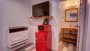 Habitación pequeña con aire acondicionado blanco, microondas rojo y televisor en un estante. Se observa un tocador de madera con espejo y lavabo al fondo.