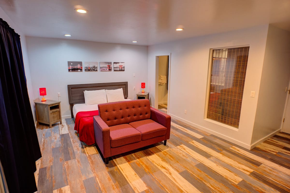 Una habitación de hotel con cama, sofá rojo, piso de madera, mesita de noche con lámpara y una pared decorada con cuadros.