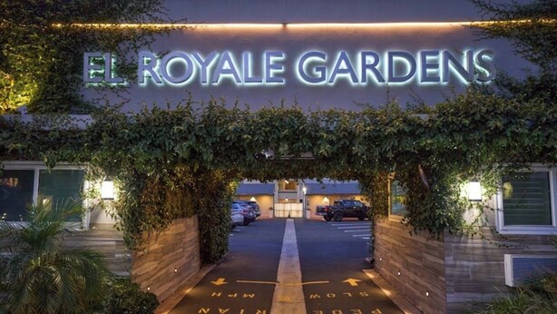 De ingang van El Royale Gardens met een bord boven de ingang en een parkeerplaats met pijlen.