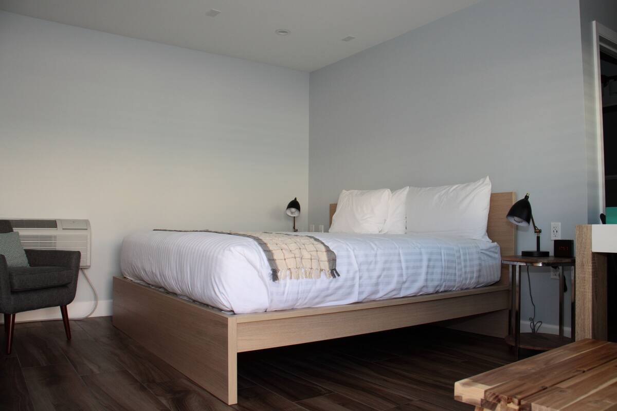 Un dormitorio con cama, mesita de noche, lámpara, silla y suelo de madera.
