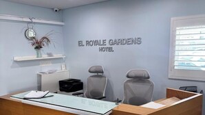 Een receptiebalie met een bord waarop 'EL ROYALE GARDENS HOTEL' staat, en twee stoelen.