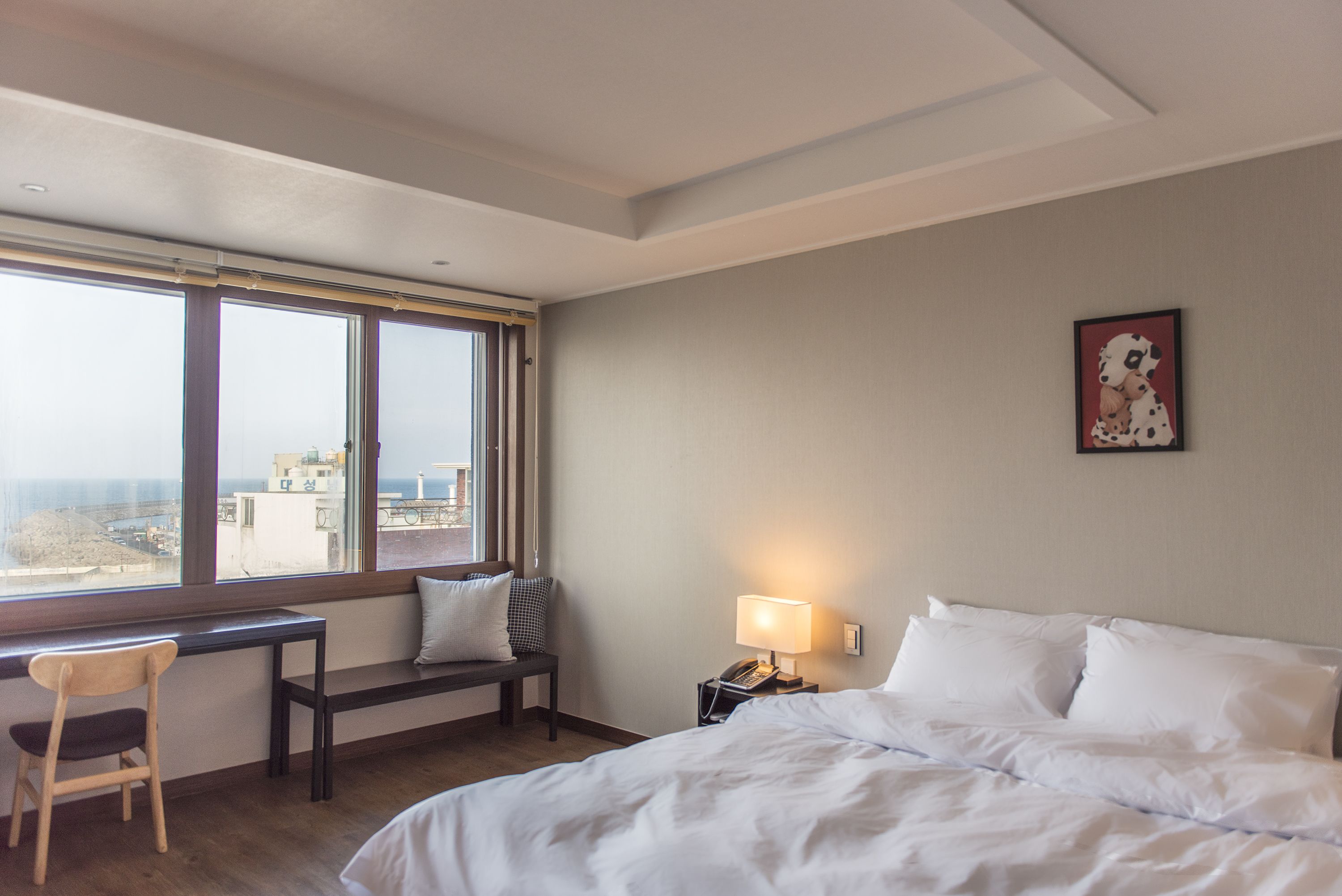 A hotel room with a bed, a desk, a chair, a nightstand with a lamp, and a window with a city view.