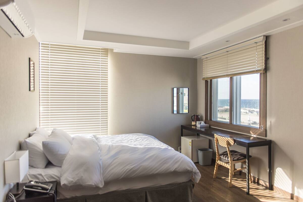 A bedroom with a bed, a desk, a chair, a window with blinds, and a view of the sea.