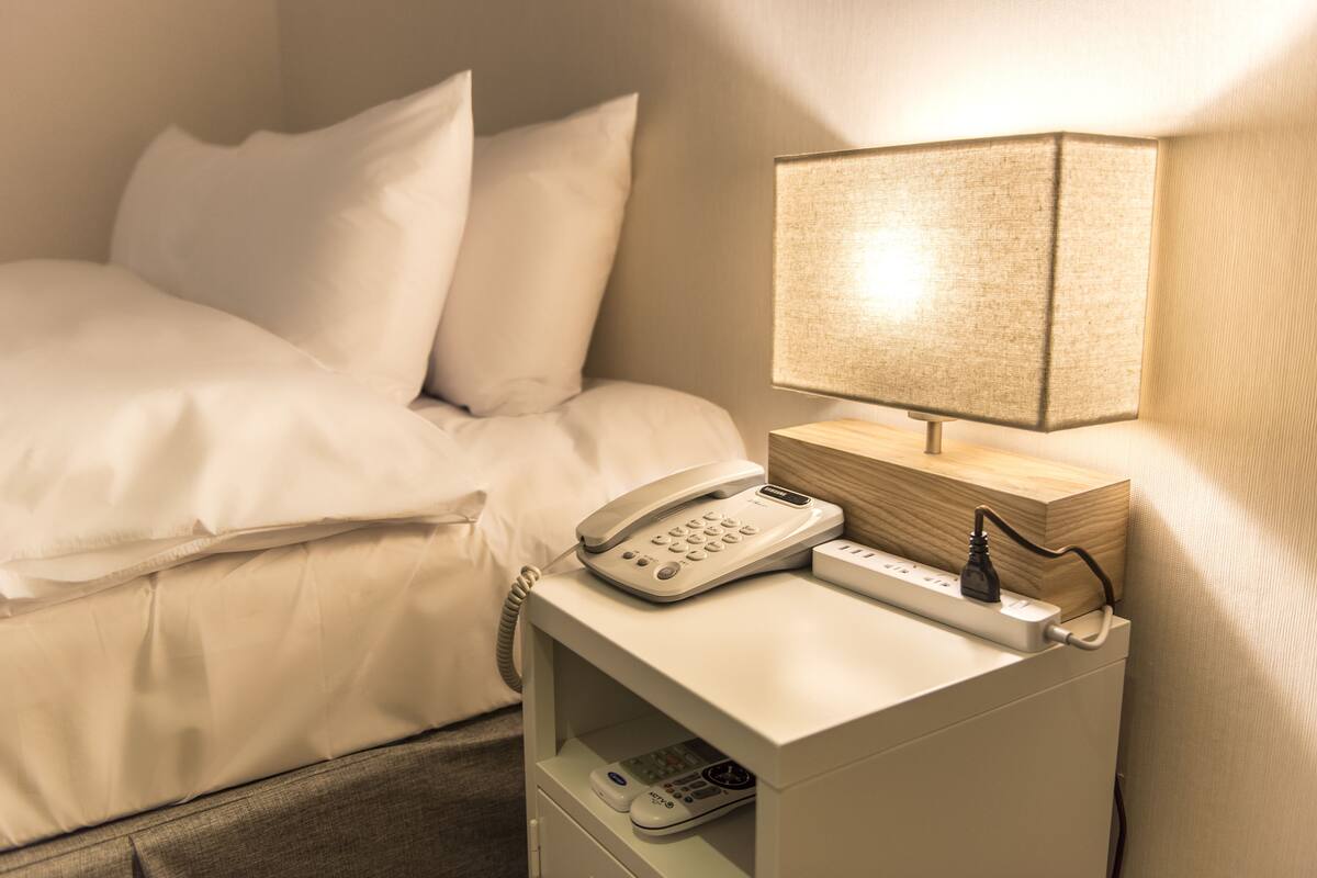 A hotel room with a bed, a bedside table with a phone and a lamp, and a power strip.