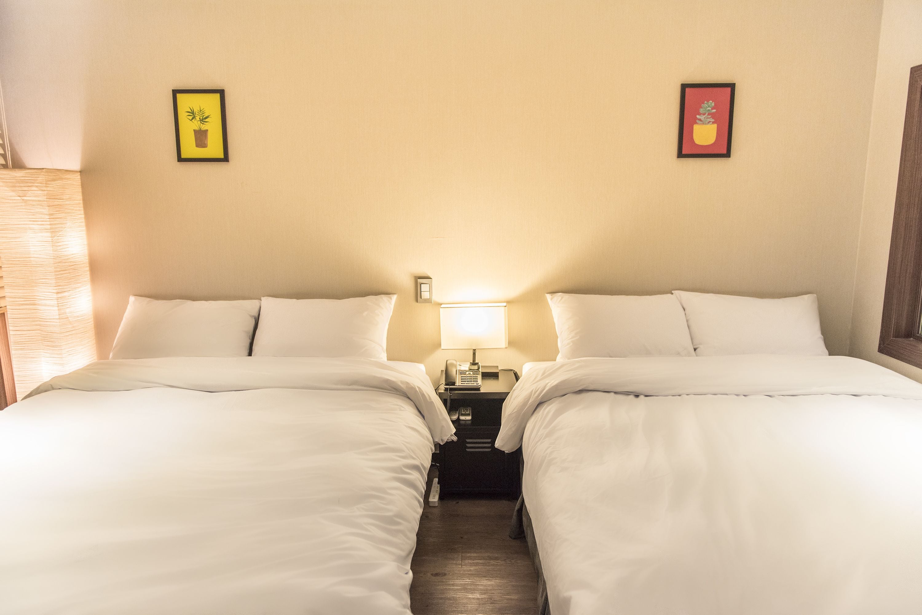 A hotel room with two beds, a nightstand with a lamp, and two framed pictures on the wall.