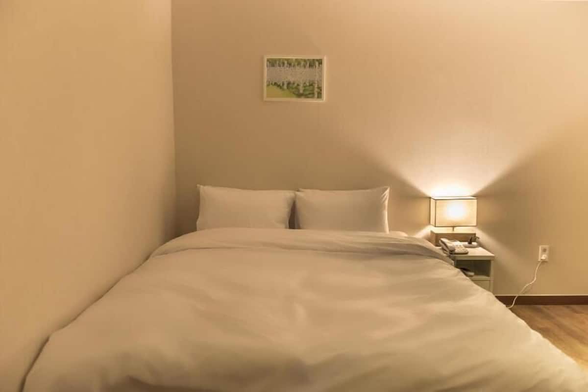 A single bed with a white comforter, a bedside table with a lamp, and a small picture frame on the wall.