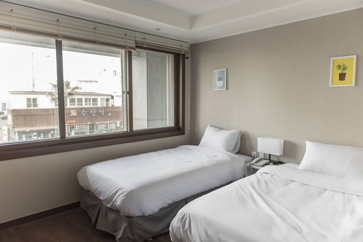 A hotel room with two single beds, a bedside table with a lamp, and a window with blinds.