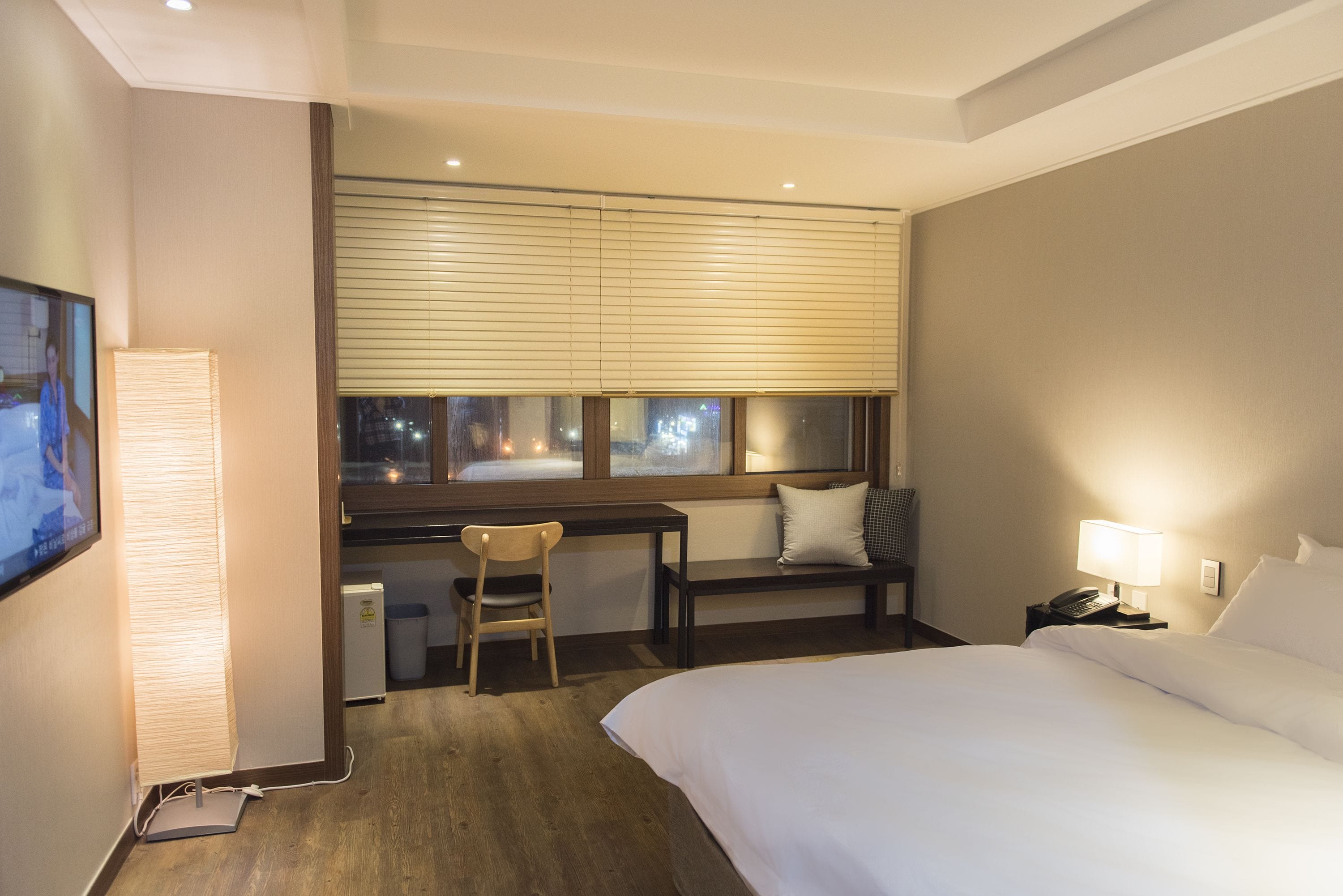 A hotel room with a bed, a desk with a chair, a window with blinds, a TV, and a lamp.