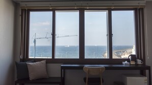 A room with a large window overlooking the sea, a desk, a chair, and a small table with a kettle and cup. - Check Inn Hotel Jeju (Jeju City)
