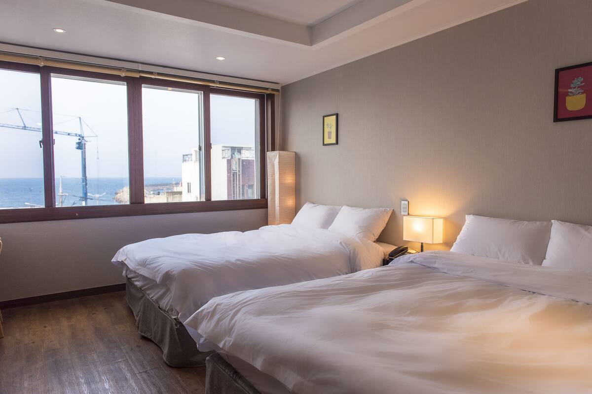 A hotel room with two beds, a large window with a view of the sea and construction cranes, and a nightstand with a lamp.