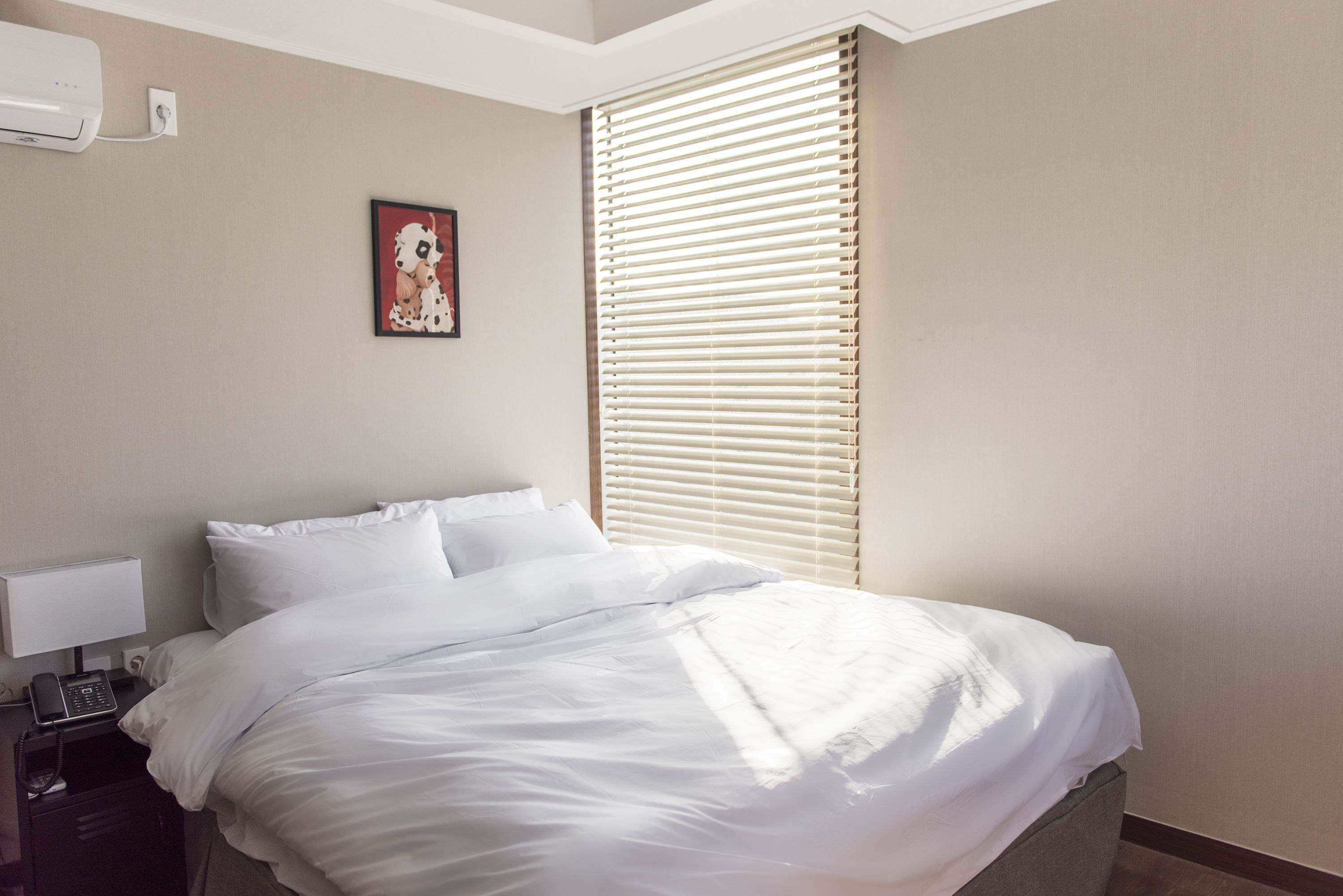 Habitación de hotel con cama, mesita de noche con lámpara, un cuadro y ventana con persianas.