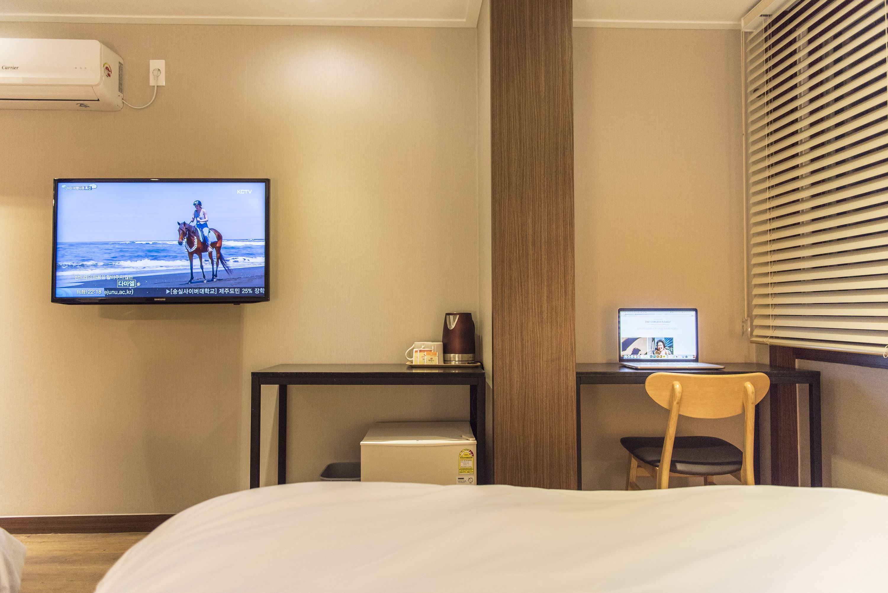 A hotel room with a bed, a TV mounted on the wall, a desk with a laptop, a chair, and a coffee maker.