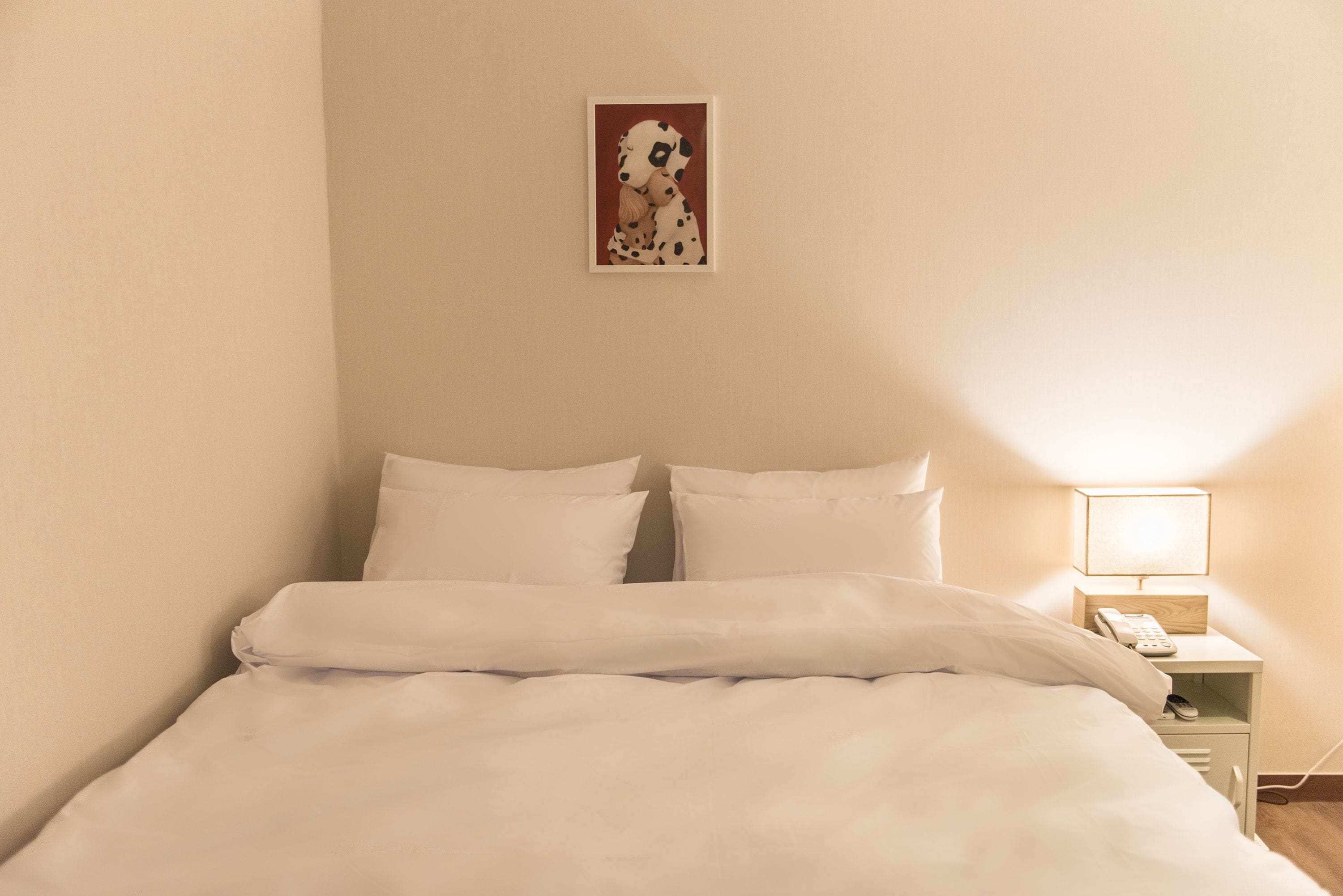 Una cama bien tendida con sábanas blancas, una mesita de noche con una lámpara y un cuadro en la pared.