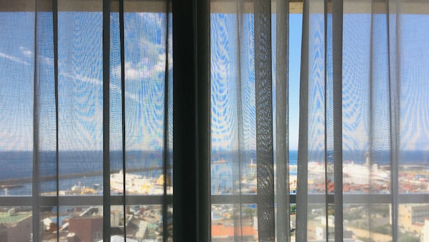 A view through a window with vertical blinds, featuring a coastal cityscape with buildings and a bridge. - Harbor Hotel (Jeju City)