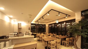 A modern dining area with wooden tables and chairs, a large potted tree, and a counter with kitchen appliances. - Harbor Hotel (Jeju City)