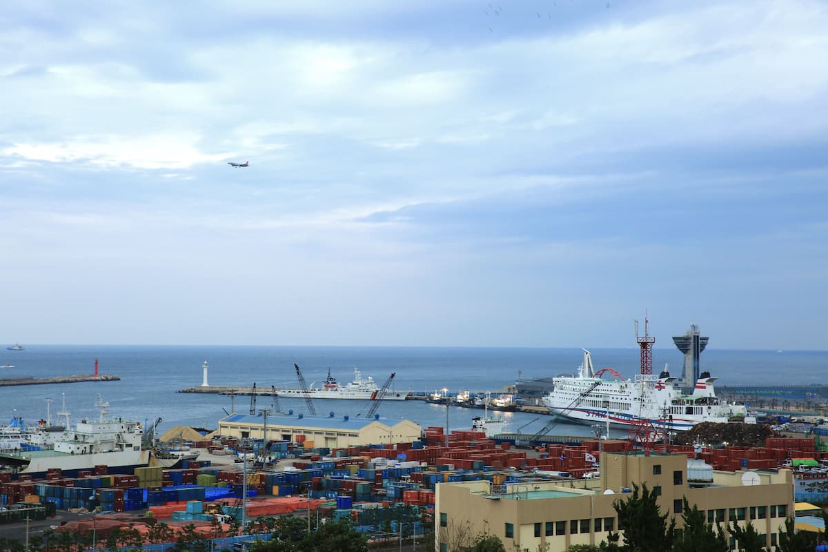 A port with shipping containers, a large ship, and a lighthouse in the background.