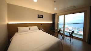A hotel room with a large bed, a desk with a chair, a view of the city, and a window with curtains. - Harbor Hotel (Jeju City)