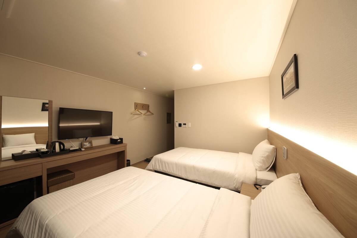 A hotel room with two beds, a desk with a flat-screen TV, a mirror, and a wall-mounted light.