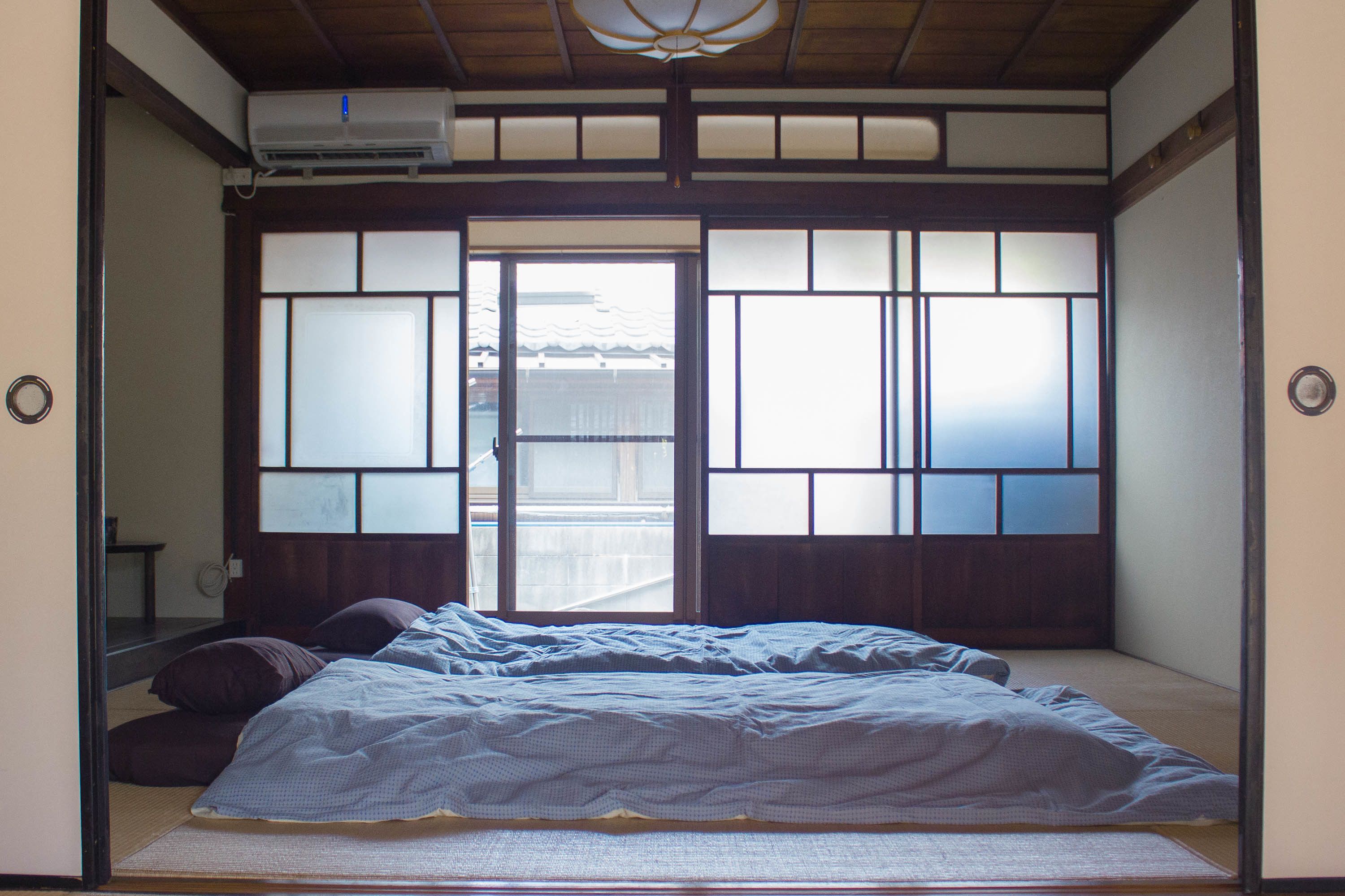 Une pièce de style japonais traditionnel, avec un sol recouvert de tatamis, un futon servant de lit, des portes coulissantes en papier de riz (shoji) et une vue sur un bâtiment traditionnel situé à l’extérieur.
