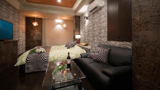 Ein Zimmer mit einer Wand im Zebra-Muster, einem Bett mit einer grün-weißen Tagesdecke, einer schwarzen Ledercouch, einem Glastisch mit Wein und Blumen sowie einem Fernseher auf der linken Seite.