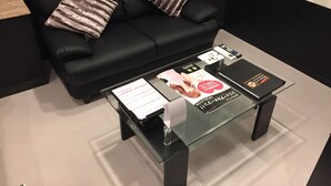 Ein modernes Wohnzimmer mit einem schwarzen Ledersofa, zwei gestreiften Kissen, einem Glastisch mit Zeitschriften und einer Fernbedienung sowie einem kleinen Beistelltisch.