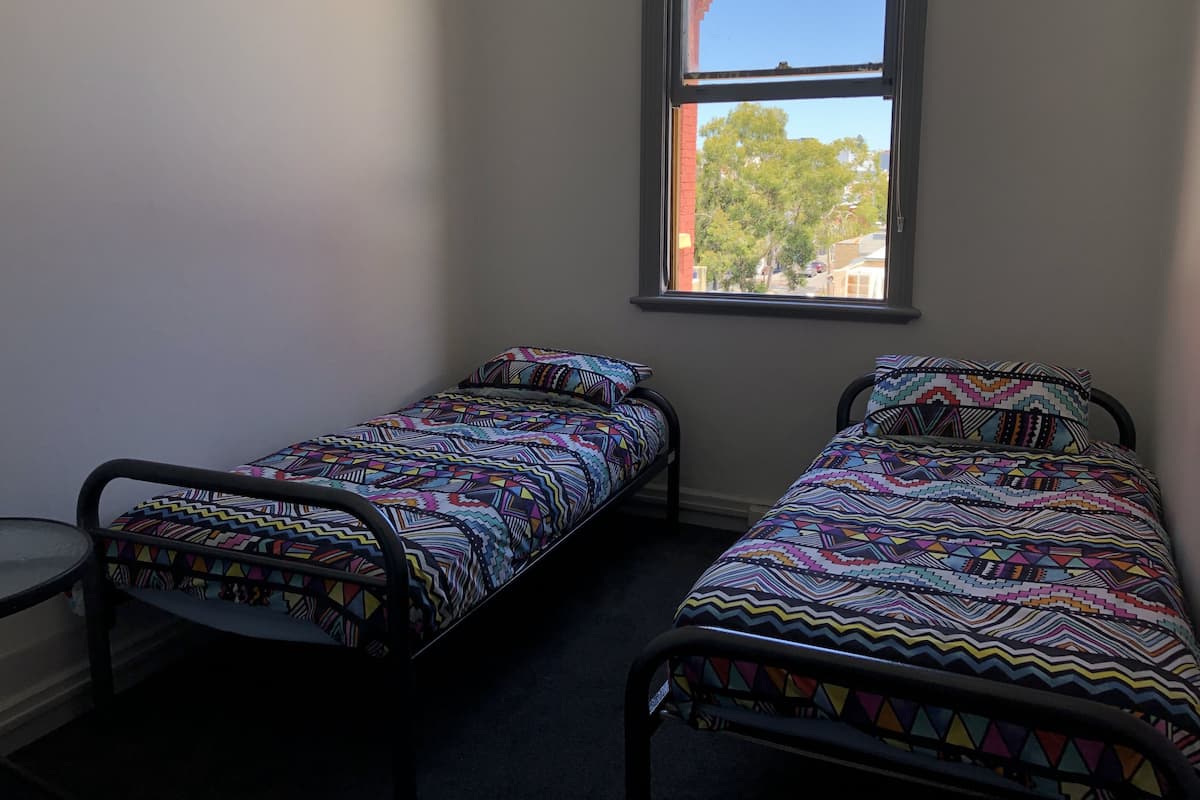 Ein kleines Zimmer mit zwei Einzelbetten, farbenfroher Bettwäsche, einem kleinen runden Tisch und einem Fenster mit Blick auf Bäume und Gebäude.