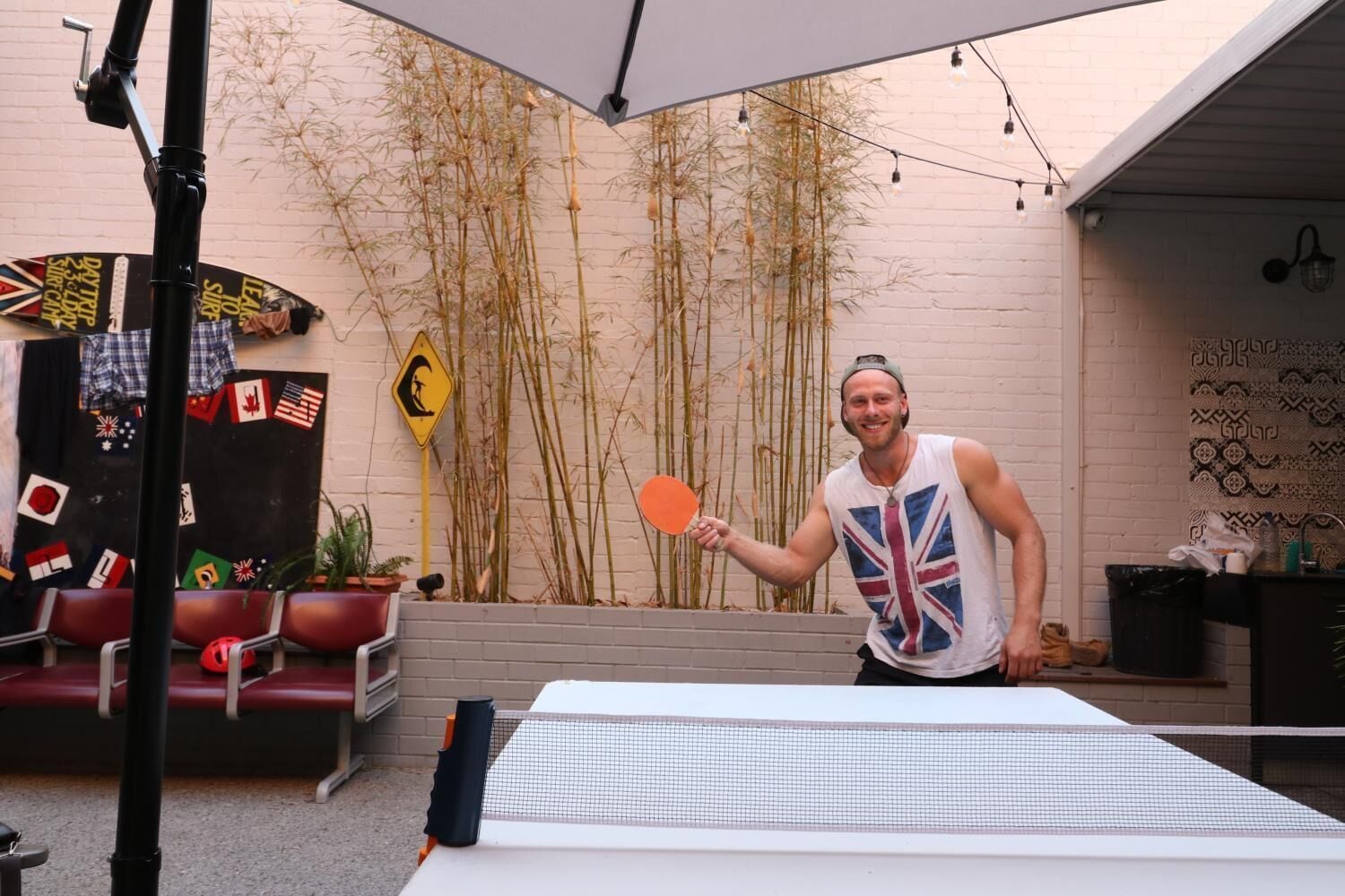 A man playing table tennis in an outdoor setting with a white wall, bamboo plants, and a table tennis table.