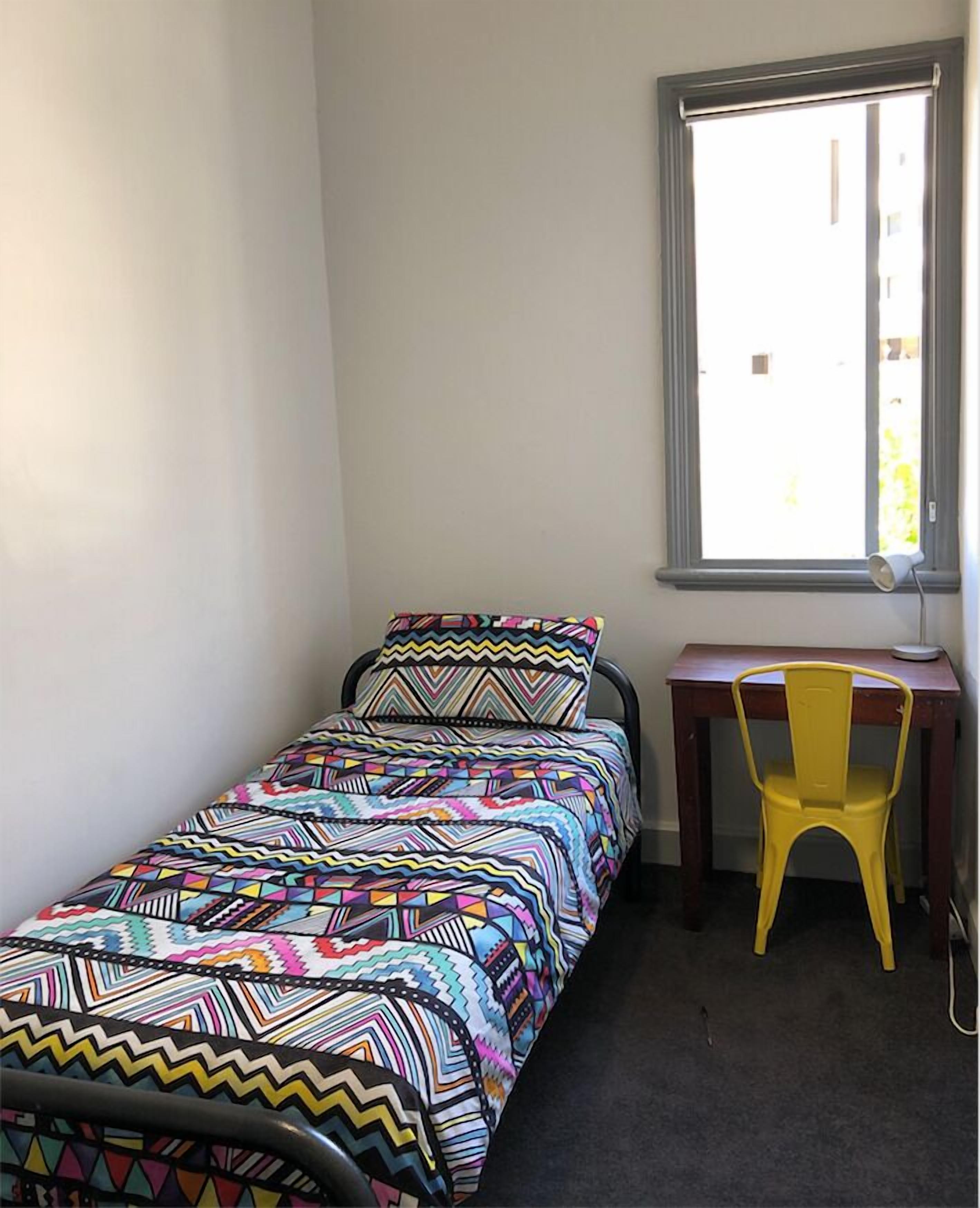 A small, single-bed room with a colorful bedspread, a wooden desk, and a yellow chair.