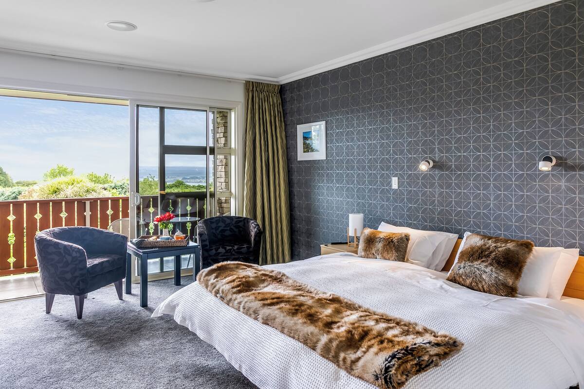 A bedroom with a large bed, a chair, a table, and a view of the outdoors.