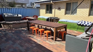 A patio with a wooden table and orange stools, a brick wall, and a green hedge.