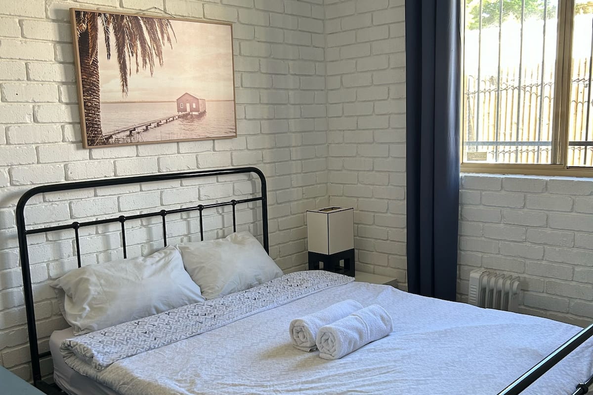 A neatly made bed with white linens and rolled towels, a white brick wall, a ceiling fan, and a window with a view of bamboo outside.