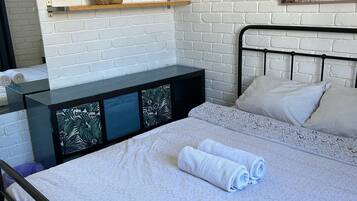 Een net opgemaakt bed met witte lakens en opgerolde handdoeken, een zwart metalen hoofdbord, een witte bakstenen muur, een klein plankje met een plantje en een ingelijste foto aan de muur.