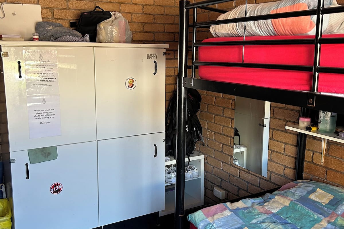 A dormitory room with bunk beds, a white wardrobe, a ceiling fan, and a brick wall.