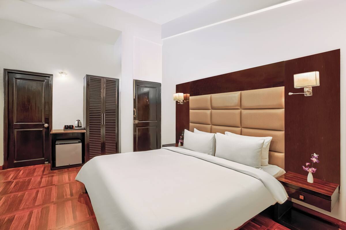 A hotel room with a large bed, wooden wardrobe, bedside table, and a small desk.
